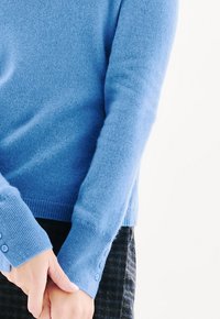 Helles blaues Sweater aus weichem Strickmaterial, mit gerippten Bündchen und Knopfdetails. Nahaufnahme von Händen, die sich in der Taille gefaltet sind.