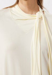 Primer plano de una blusa de crema que presenta un detalles de una bufanda anudada en el escote, llevada por una persona con cabello largo castaño.