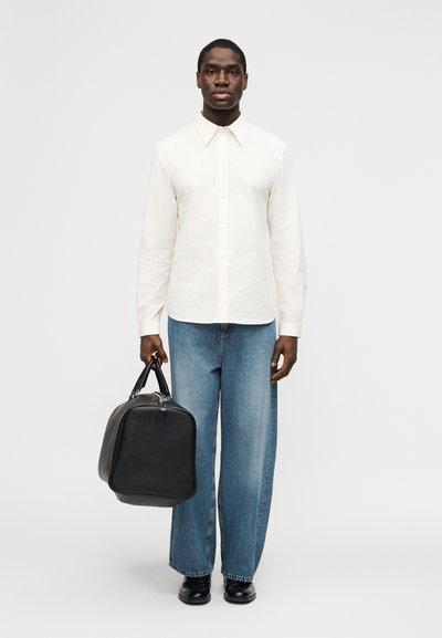 Uomo con camicia bianca a bottoni e jeans blu a gamba larga, che tiene una borsa nera in pelle, in piedi davanti a uno sfondo grigio semplice.