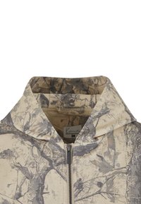 Beige väritetty huppari, jossa on maastokuvio. Etuosassa vetoketju, teksturoitu kangas ja brändilogot vetoketjun läheisyydessä.