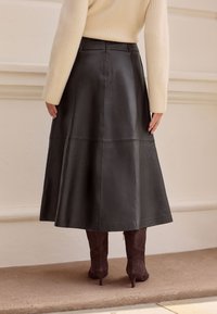 Schwarzer Leder-Midirock mit ausgestelltem Design, glatter Textur und Nahtdetails, kombiniert mit braunen Wildleder-Stiefeletten mit Absatz.