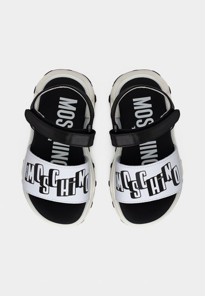 Ζεύγος μαύρων και λευκών σανδαλιών Moschino με ρυθμιζόμενους ιμάντες και έντονη ετικέτα με επωνυμία στα μπροστινά λουριά.