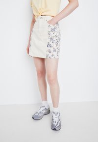 Beige denimskjørt med blomstertrykk på sidene, med høy midje, frontlommer og knappelukking. Brukt sammen med grå treningssko.