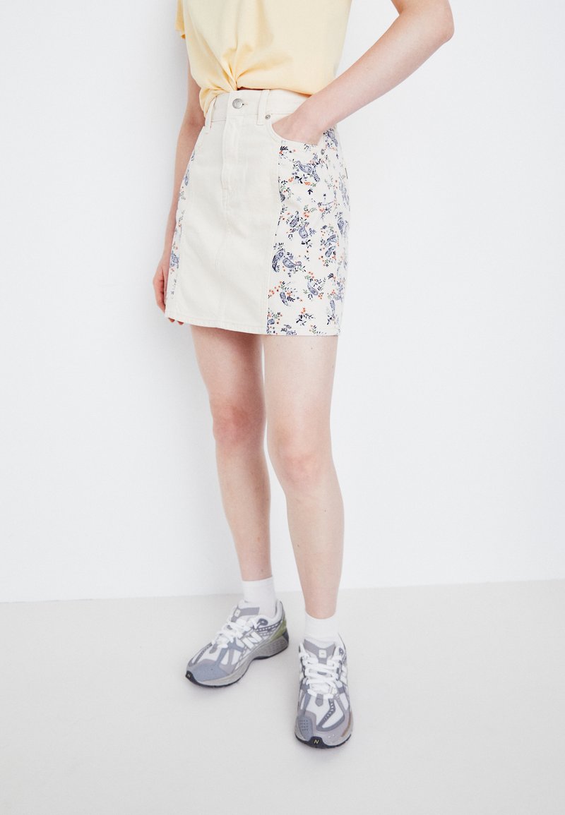 Beige denimskjørt med blomstertrykk på sidene, med høy midje, frontlommer og knappelukking. Brukt sammen med grå treningssko.
