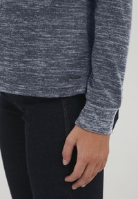 Mørkeblå melert langermet topp med rund nederkant og ribbede mansjetter, kombinert med mørke jeans. Merket er synlig nederst til venstre.