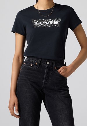 Person iført sort Levi's t-shirt med hvidt logo og sorte højtaljede jeans, den ene hånd i lommen, foran ensfarvet grå baggrund.
