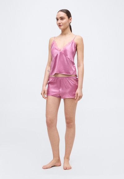 Γυναίκα που στέκεται ξυπόλυτη φορώντας ροζ σατινέ καμεζόλη με δαντελωτό τελείωμα και ταίριαζα σορτς, μπροστά σε απλό φωτεινό φόντο.