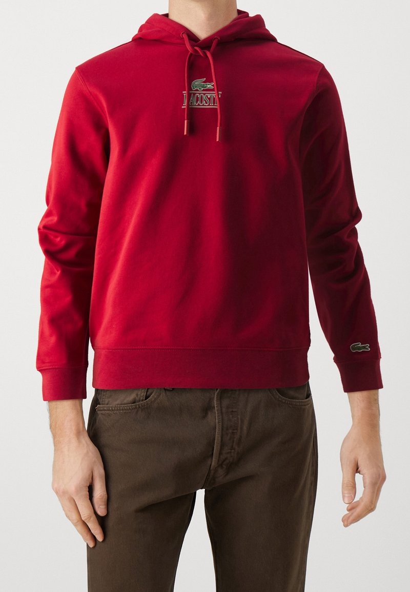 Homme portant un sweat à capuche rouge avec le logo Lacoste sur la poitrine et la manche, associé à un pantalon marron, debout devant un fond uni.