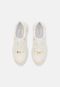 Sneakers bianchi in pelle con texture trapuntata, dettagli dorati e lacci. Presentano un logo sulla linguetta e un design della punta arrotondata.