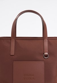 Bolso tote marrón con una superficie texturizada, asas superiores dobles y un bolsillo frontal deslizante con marca en relieve. Acentos suaves y herrajes circulares.