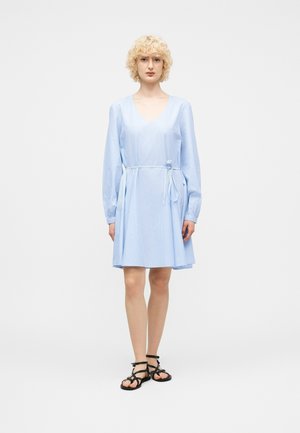 Osoba s krátkymi kučeravými blond vlasmi, oblečená v svetlomodrých šatách s dlhým rukávom, zviazaných v páse, a čiernych sandáloch na remienky, stojaca na bielom pozadí.