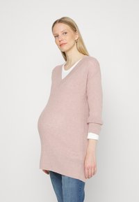 Sweater rosa claro de mangas longas, com decote em V e textura canelada. Usado em camadas sobre uma blusa branca. Corte solto, projetado para acomodar uma silhueta grávida.