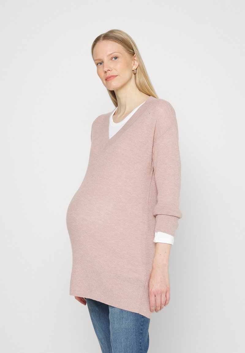 Sweater rosa claro de mangas longas, com decote em V e textura canelada. Usado em camadas sobre uma blusa branca. Corte solto, projetado para acomodar uma silhueta grávida.