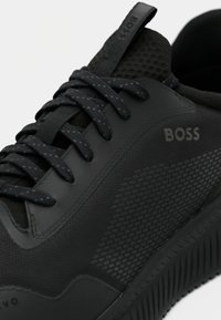 Μαύρο αθλητικό παπούτσι με υφή, που διαθέτει λεία επιφάνεια, μαύρα κορδόνια και λεπτομέρεια με λογότυπο στο πλάι. Εξωτερική σόλα από καουτσούκ με πρόσφυση.