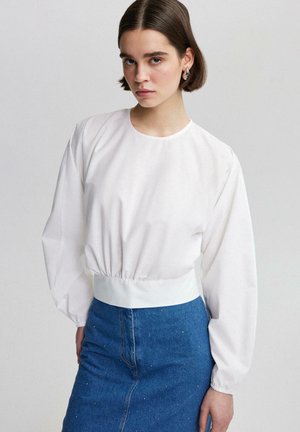Eine Frau mit kurzen braunen Haaren trägt eine langärmlige weiße Bluse mit taillierter Passform und einen blauen Denim-Rock mit kleinen Nieten.