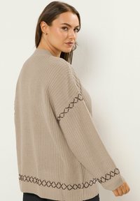 Vaaleanbeige neulecardigan, jossa on tekstuuripinta ja ruskeita brodeerattuja yksityiskohtia hihansuissa ja helmoissa. Rentoa mallia ja pyöreä pääntie.