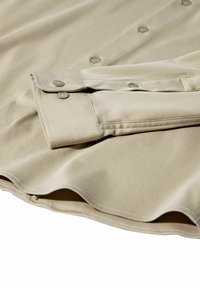 Bežinė ilgomis rankovėmis marškiniai su mygtukų užsegimais, mygtukų eilių ir matoma siuvimo linija minkšto audinio fone.