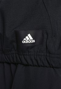 Schwarze Sportjacke aus strukturiertem Stoff, mit elastischem Bund und einem rechteckigen Adidas-Logo-Patch in Weiß.