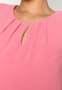 Blusa rosa con scollatura rotonda e aperture a forma di chiave, dettagli plissettati e una texture di tessuto morbido e leggero.