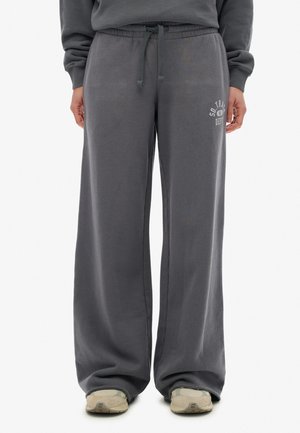 Personne portant un pantalon de survêtement gris ample avec taille à cordon et des baskets beiges, se tenant contre un fond blanc.