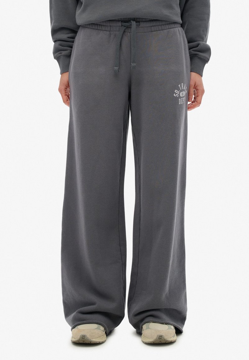 Person in lockeren grauen Sweatpants mit Kordelzug und beigen Turnschuhen, der vor einem weißen Hintergrund steht.