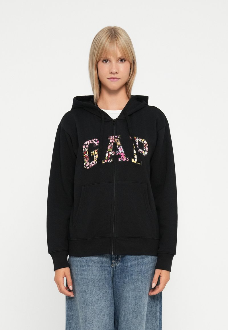 Felpa nera con zip caratterizzata da un logo floreale "GAP", due tasche frontali e cappuccio con coulisse. Tessuto morbido e di peso medio.