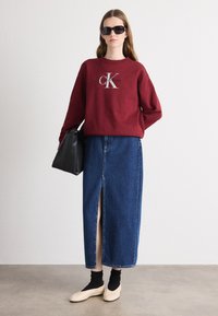 Felpa bordeaux con logo 'Calvin Klein', abbinata a una gonna di jeans blu con uno spacco frontale. Borsa nera, calze nere e scarpe beige.