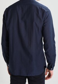 Maglietta a maniche lunghe blu navy con una texture sottile, polsini a bottoni e colletto standard. Presenta un orlo curvo e una finitura del tessuto liscia.