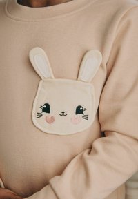 Light pink sweatshirt with a fluffy beige bunny face patch featuring embroidered eyes and rosy cheeks. Soft texture, round neckline.