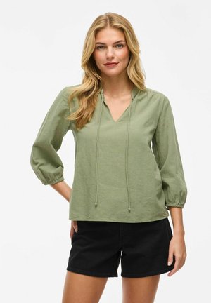 Haut vert clair à manches trois-quarts avec un décolleté en V et des détails de lien ajustables. Tissu doux et texturé, associé à un short noir.