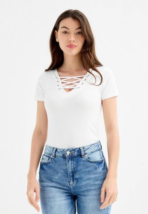 Mujer con cabello largo y castaño lleva una camiseta blanca de manga corta con detalle de encaje cruzado y vaqueros azules de tiro alto, de pie sobre un fondo blanco.