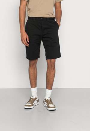 Schwarze Shorts mit maßgeschneidertem Design, mit flacher Vorderseite, seitlichen Taschen und umgeschlagenen Säumen. Kombiniert mit weißen Kurzsocken und braunen Sneakers.