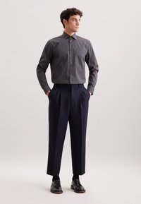 Camicia button-up blu navy con motivo geometrico bianco, abbinata a pantaloni plissettati blu navy e scarpe nere stringate. Tessuto liscio, vestibilità su misura.