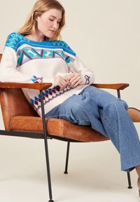 Weißer Strickpulli mit einem farbigen geometrischen Muster an den Schultern und Manschetten, kombiniert mit hellblauen, weit geschnittenen Jeans. Model sitzt in einem Stuhl.