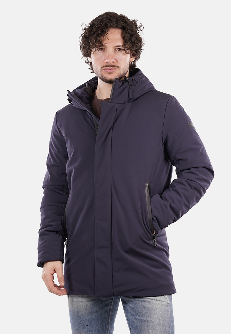 Uomo con capelli ricci che indossa un giubbotto invernale con cappuccio di colore viola scuro e jeans chiari strappati, in piedi contro uno sfondo bianco.