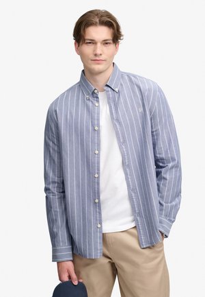 Ung mand iført en blå og hvid stribet skjorte med knapper over en hvid t-shirt og beige bukser, holder en mørkeblå kasket i venstre hånd.