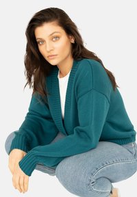 Turkusowy sweter dziergany z prążkowanymi mankietami i otwartym przodem, noszony na białej koszulce. Model ma rozczochrane włosy i siedzi na neutralnym tle.