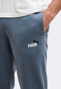 Άνδρας που φοράει μπλε αθλητική φόρμα Puma με το χέρι στην τσέπη και μέρος λευκού πουκάμισου ορατό πάνω από τη μέση.
