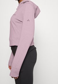 Světle fialová mikina s kapucí a texturovaným vzorem. Má dlouhé rukávy s otvory pro palce; doplněná černými kalhotami se zipy.