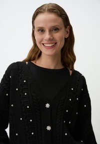 Zwarte gebreide cardigan met een V-hals, een gestructureerd kabelpatroon en decoratieve faux edelstenen langs de voorkant. Zacht, comfortabel materiaal.