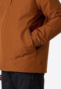 Chaqueta marrón impermeable con cierre de cremallera, bolsillos laterales y un corte relajado. Presenta la marca "DRYVENT" en la manga, textura suave.