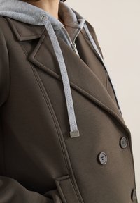 Gros plan d'un manteau marron à double boutonnage porté par-dessus un sweat à capuche zippé gris avec cordons blancs comportant un texte en relief.