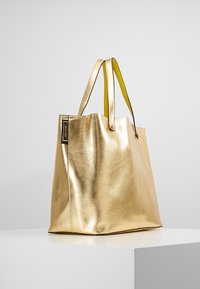 Zlatá metalická taška přes rameno s hladkým, jemným povrchem. Má žluté popruhy a černou cedulku na boku. Obdélníkový tvar, otvorený design na vrchu.