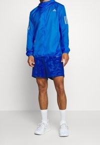 Mężczyzna ubrany w niebieską kurtkę z kapturem marki Adidas oraz dopasowane, wzorzyste spodenki, białe skarpetki i białe buty sportowe.