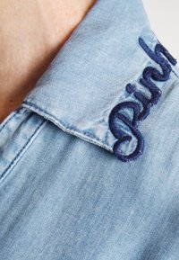 Col de chemise en denim bleu clair avec texte en cursive brodé en bleu foncé partiellement visible près de l'encolure.