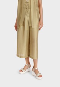 Culotte di colore kaki, morbidi e larghi, abbinati a una blusa leggera coordinata. La modella indossa sandali con piattaforma color crema con cinturini decorativi.