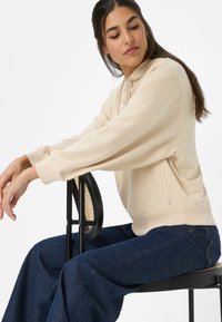 Beige sweatshirt met capuchon, geribbelde manchetten en zijslitdetails, gedragen met donkerblauwe wijde jeans, zittend op een zwarte stoel met rotanaccenten.