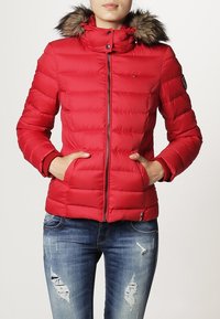 Rød puffjakke med en hætte foret med pels, lynlåslukning foran og polstrede sektioner. Har sidelommer og ribbede manchetter. Bæres med blå slidte jeans.