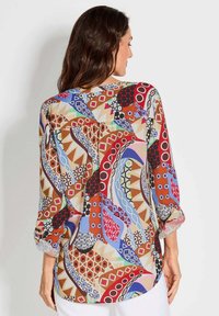 Multikleurige blouse met een mix van geometrische en abstracte patronen, semi-doorzichtige stof en driekwartmouwen met opgerolde manchetten.