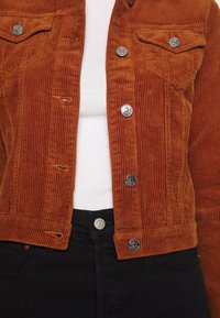 Roestkleurige corduroyjas met zilveren drukknoopjes, gedragen over een witte ribbelshirt en zwarte high-waisted broek.